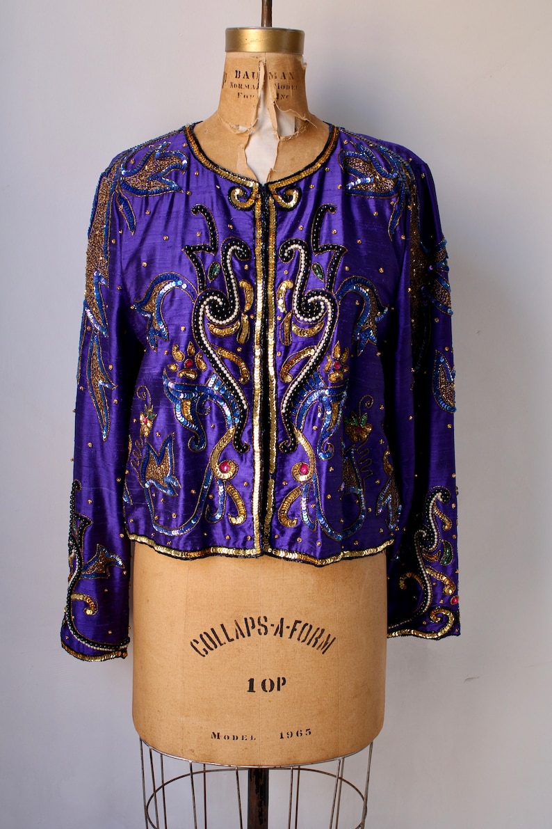 May include: A purple sequined jacket with gold and blue embroidery. The jacket has a zipper closure and long sleeves. The jacket is displayed on a mannequin with the words 'COLLAPS-A-FORM 1 OP MODEL 1965' printed on the mannequin's torso.