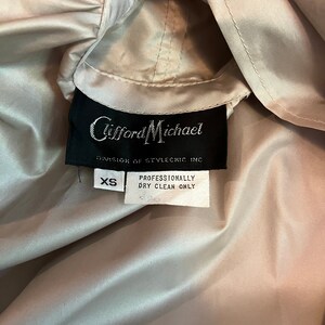 Op de afbeelding: Close-up van een lichtbeige jas met capuchon. Het zwarte label luidt "Clifford Michael, Division of Stylechic Inc." en bevat een label met de tekst "XS" en "Professioneel chemisch reinigen alleen".