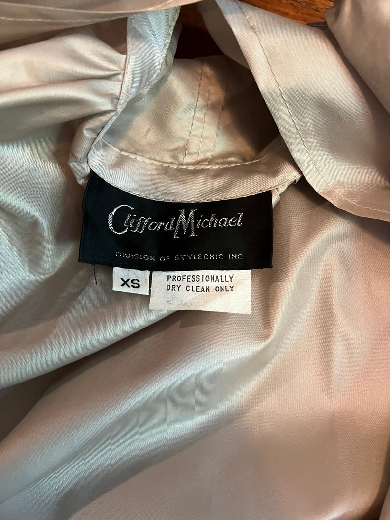 Op de afbeelding: Close-up van een lichtbeige jas met capuchon. Het zwarte label luidt "Clifford Michael, Division of Stylechic Inc." en bevat een label met de tekst "XS" en "Professioneel chemisch reinigen alleen".