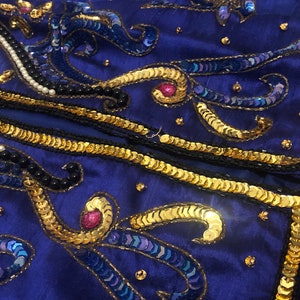 May include: Close-up of a section of a royal blue fabric, heavily embellished with gold sequins, black and white beads, and iridescent blue sequins. The design features swirling patterns and borders, creating a luxurious and ornate appearance. The fabric is likely part of a garment.