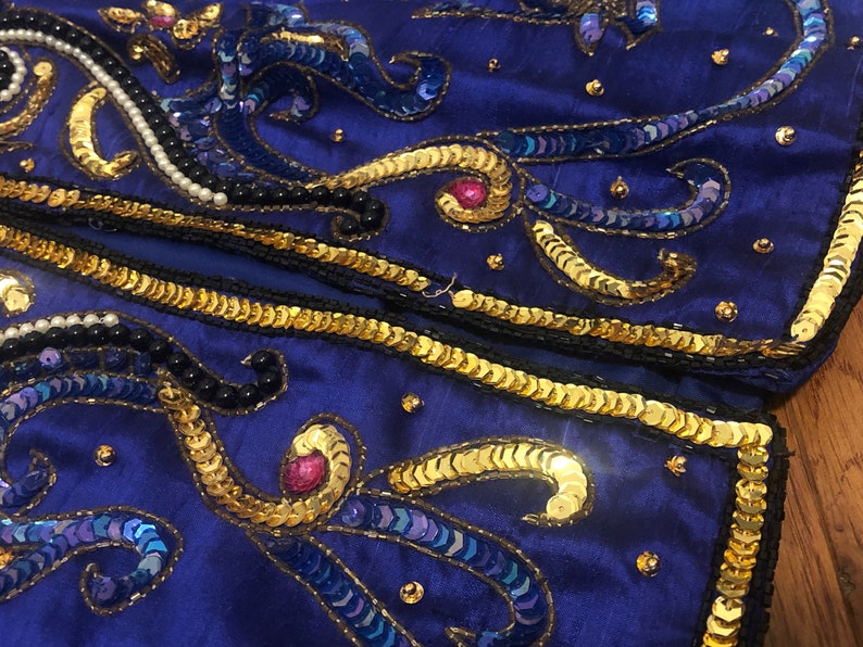 May include: Close-up of a section of a royal blue fabric, heavily embellished with gold sequins, black and white beads, and iridescent blue sequins. The design features swirling patterns and borders, creating a luxurious and ornate appearance. The fabric is likely part of a garment.