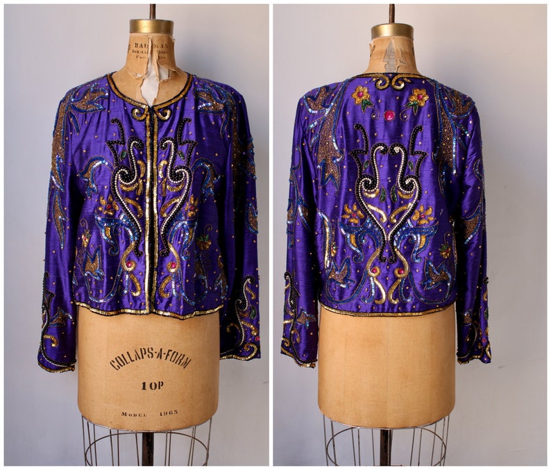 May include: A vintage purple jacket with intricate beadwork and sequin embellishments. The jacket features a round neckline, long sleeves, and a cropped silhouette. The design includes gold, black, and blue accents, creating a decorative pattern. The jacket is displayed on a vintage dress form.