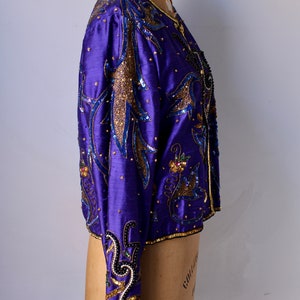 May include: A vibrant purple jacket with intricate gold and blue sequin and bead embellishments. The jacket features long sleeves and a round neckline, displayed on a mannequin. The design includes floral and abstract patterns.