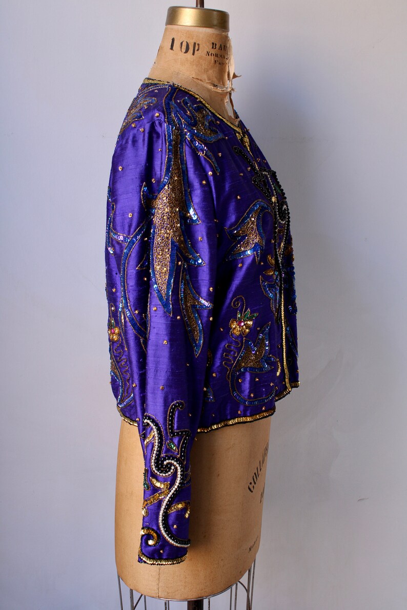 May include: A vibrant purple jacket with intricate gold and blue sequin and bead embellishments. The jacket features long sleeves and a round neckline, displayed on a mannequin. The design includes floral and abstract patterns.