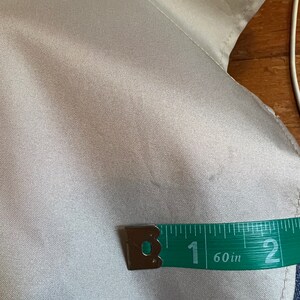 Op de afbeelding: Close-up van een licht zilverkleurige stof met een groene meetlint. De tape toont een meting van 2,54 cm. De stof lijkt een satijn- of zijdemateriaal te zijn.