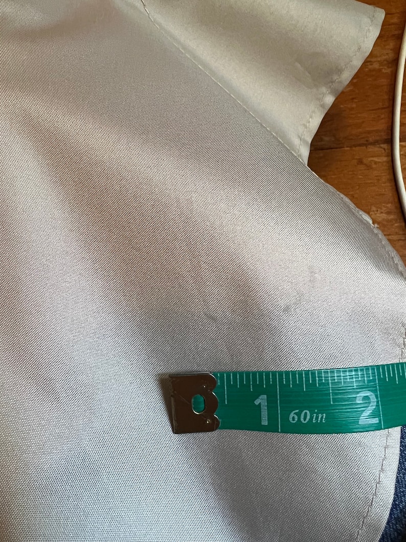 Op de afbeelding: Close-up van een licht zilverkleurige stof met een groene meetlint. De tape toont een meting van 2,54 cm. De stof lijkt een satijn- of zijdemateriaal te zijn.