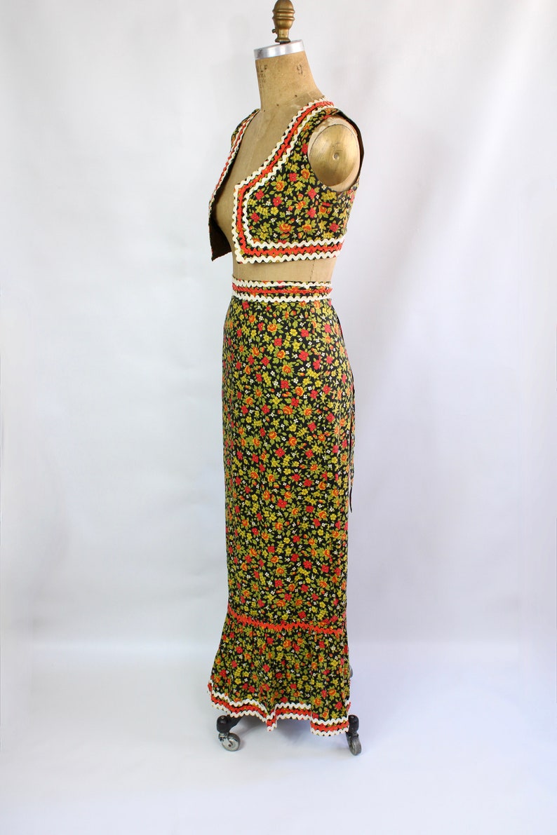 May include: A vintage two-piece outfit featuring a vest and a long skirt. The set is adorned with a floral pattern in red, yellow, and green on a black background. The edges are trimmed with white and orange lace.