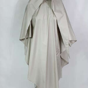 Op de afbeelding: Een lichtbeige cape met capuchon, getoond op een draadmodel. De cape heeft een vloeiend ontwerp met wijde mouwen en een gedrapeerde capuchon. De stof lijkt een lichtgewicht materiaal te zijn, geschikt voor layering. Ongeveer 150 cm lang.
