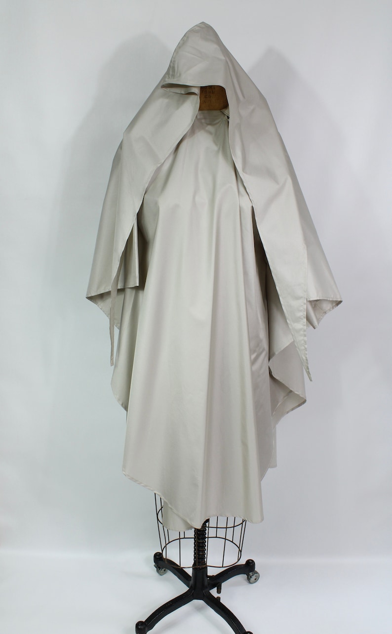 Op de afbeelding: Een lichtbeige cape met capuchon, getoond op een draadmodel. De cape heeft een vloeiend ontwerp met wijde mouwen en een gedrapeerde capuchon. De stof lijkt een lichtgewicht materiaal te zijn, geschikt voor layering. Ongeveer 150 cm lang.