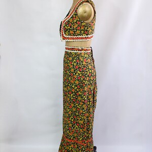May include: A two-piece floral outfit featuring a cropped vest and a long skirt. The black fabric is covered in small yellow and red flowers. The edges of the vest and skirt are trimmed with white, orange, and black decorative trim. The outfit is displayed on a mannequin.