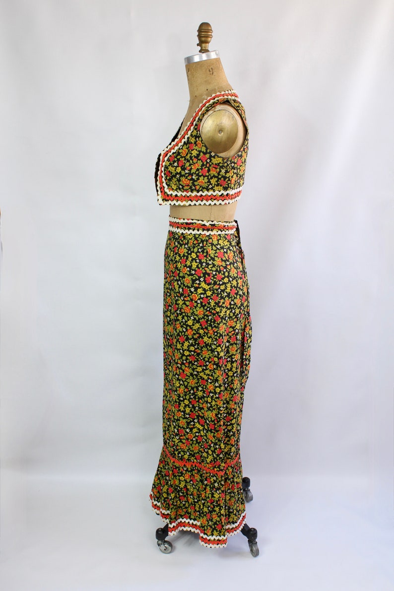 May include: A two-piece floral outfit featuring a cropped vest and a long skirt. The black fabric is covered in small yellow and red flowers. The edges of the vest and skirt are trimmed with white, orange, and black decorative trim. The outfit is displayed on a mannequin.