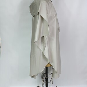 Op de afbeelding: Een lichtbeige poncho met een capuchon, weergegeven op een mannequin. De poncho heeft een vloeiend, asymmetrisch ontwerp met wijde mouwen en een gedrapeerde voorkant. De stof lijkt lichtgewicht en geschikt om in laagjes te dragen.