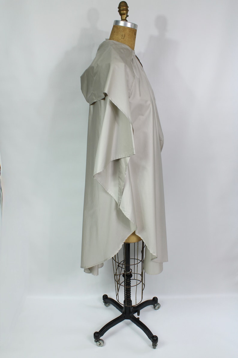 Op de afbeelding: Een lichtbeige poncho met een capuchon, weergegeven op een mannequin. De poncho heeft een vloeiend, asymmetrisch ontwerp met wijde mouwen en een gedrapeerde voorkant. De stof lijkt lichtgewicht en geschikt om in laagjes te dragen.
