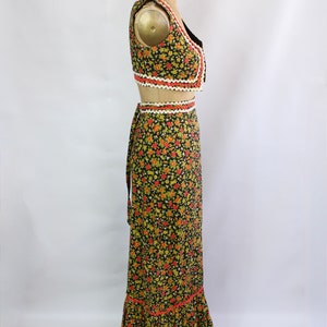 May include: A two-piece floral print outfit on a mannequin. The set includes a cropped vest and a long skirt. The black fabric is covered in small red, yellow, and green flowers. The edges are trimmed with white and orange decorative trim.