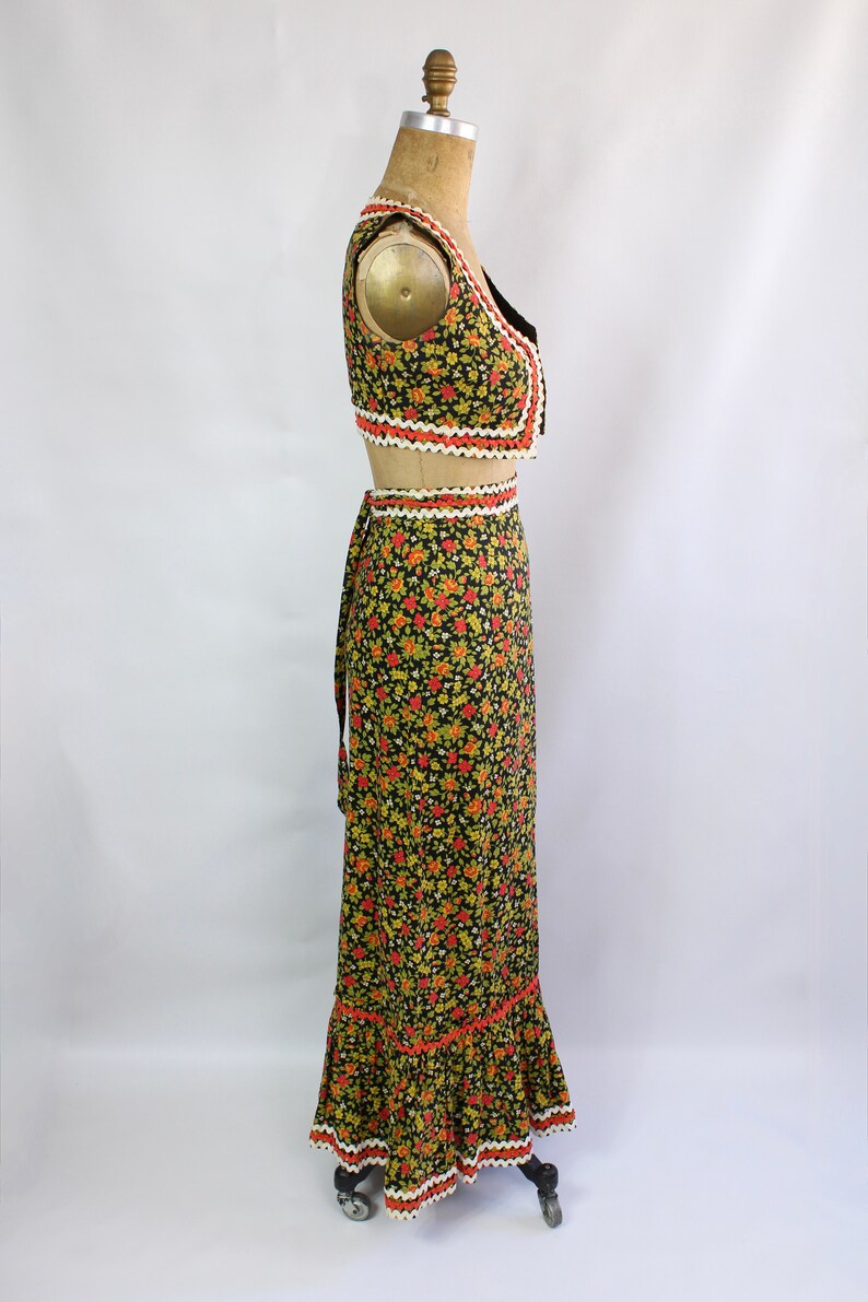 May include: A two-piece floral print outfit on a mannequin. The set includes a cropped vest and a long skirt. The black fabric is covered in small red, yellow, and green flowers. The edges are trimmed with white and orange decorative trim.