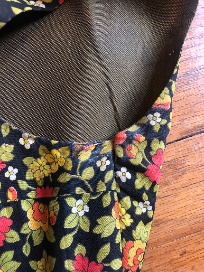 May include: Close-up of a black floral print fabric with yellow, orange, and white flowers and green leaves. The fabric has a dark background and a sheer, olive-green lining. The edges of the fabric are visible.