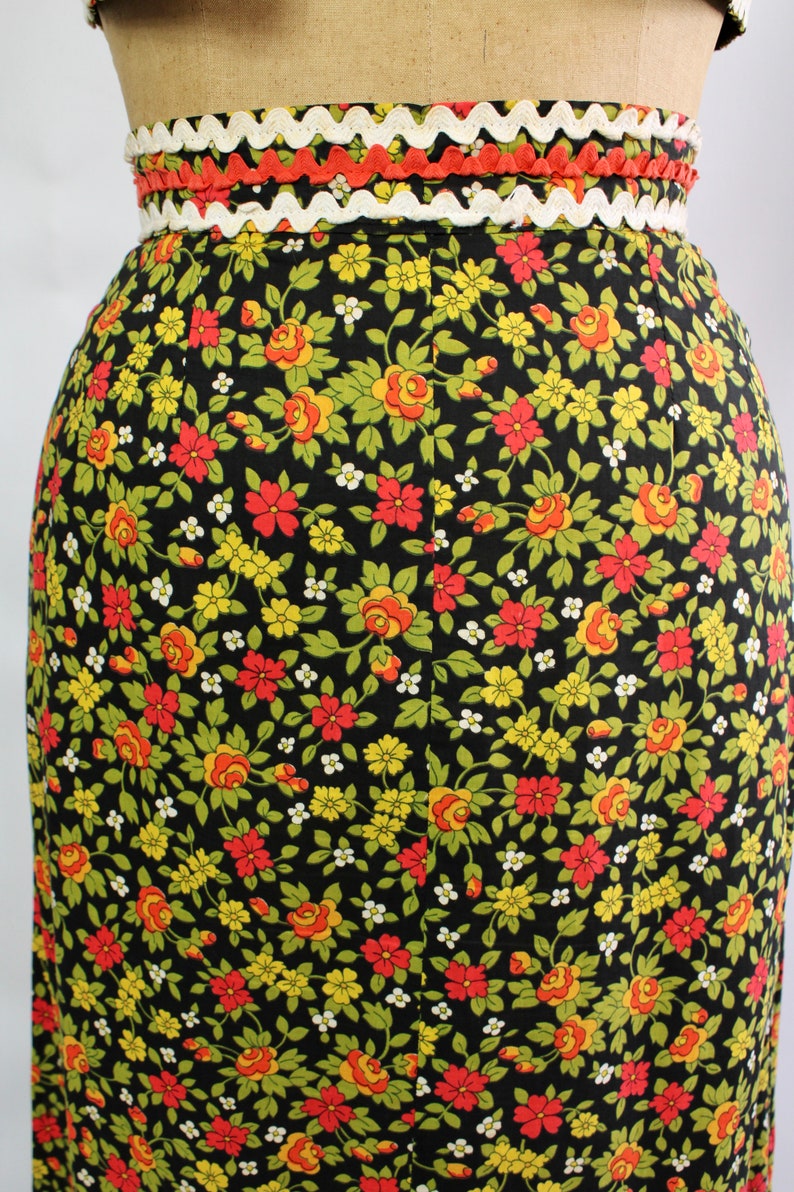 May include: A black skirt with a floral pattern featuring red, yellow, and green flowers. The waistband is trimmed with white and orange decorative trim. The skirt is a vintage style.