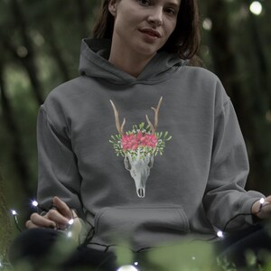 Könnte beinhalten: Ein grauer Kapuzenpullover mit einem Design eines Hirschschädels, verziert mit rosa Blumen und Geweih. Der Hoodie hat eine Vordertasche und eine Kapuze. Das Design ist auf der Brust zentriert.