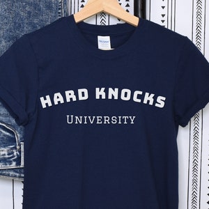 Könnte beinhalten: Marineblaues T-Shirt mit dem weißen Text "HARD KNOCKS UNIVERSITY". Das T-Shirt hängt an einem Holzbügel. Eine blaue Jeans und eine schwarz-weiß gemusterte Wand sind im Hintergrund zu sehen.