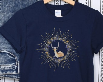 Könnte beinhalten: Marineblaues T-Shirt mit einem Hirsch-Design. Die Illustration zeigt einen Hirsch in einem kreisförmigen Rahmen, verziert mit goldenen und blauen Details. Das T-Shirt hängt an einem Holzbügel, im Hintergrund ist eine blaue Jeans zu sehen.
