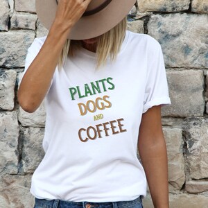 Op de afbeelding: Wit T-shirt met de woorden "PLANTS DOGS AND COFFEE" in groen, oranje en bruin. Het shirt wordt gedragen met een blauwe spijkerbroek en een bruine hoed. De achtergrond is een stenen muur.