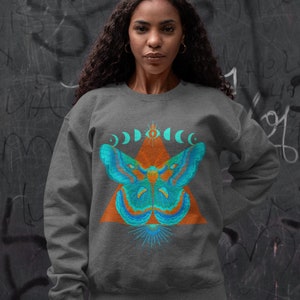 May include: Dark gray sweatshirt featuring a teal and orange moth design. The moth is centered on an orange triangle, with a series of crescent moons above. A sunburst design is below the moth.
