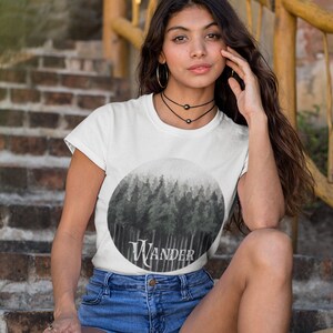 May include: White t-shirt with a circular graphic of a forest scene in shades of gray and the word "Wander" in white script. The model is wearing denim shorts and a black choker necklace.