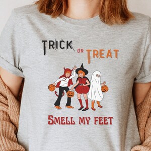 May include: A light gray t-shirt with the words "Trick or Treat" and "Smell My Feet" printed on it. The shirt features a cartoon illustration of three children in Halloween costumes, each holding a jack-o'-lantern.