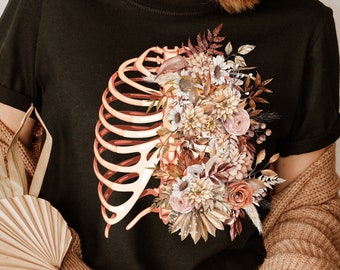 May include: Black t-shirt with a rib cage design on the left and a floral arrangement on the right. The flowers are in beige, brown, and pink. The shirt has short sleeves and a crew neck.