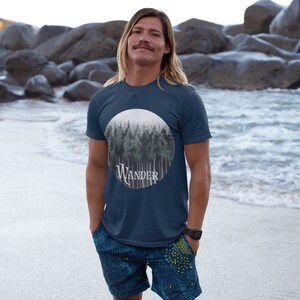 May include: Navy blue t-shirt featuring a circular graphic of a forest scene with the word "Wander" in white script. The shirt is worn with blue patterned shorts. The person is standing on a sandy beach with the ocean in the background.