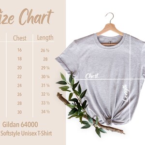May include: A light gray Gildan 64000 Adult Softstyle Unisex T-Shirt hangs on a wooden hanger. A size chart shows chest and length measurements in inches for sizes XS to 4XL. The shirt is styled with a knot at the bottom and a sprig of greenery.