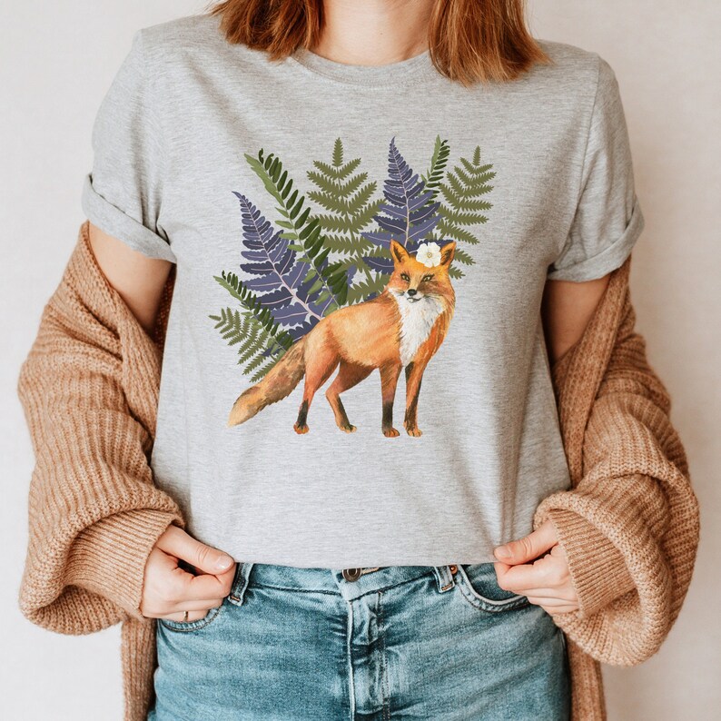 May include: A light grey t-shirt with a watercolour illustration of a fox wearing a flower crown, set against green and purple ferns. The fox has orange fur and a white chest. The shirt has rolled-up sleeves.