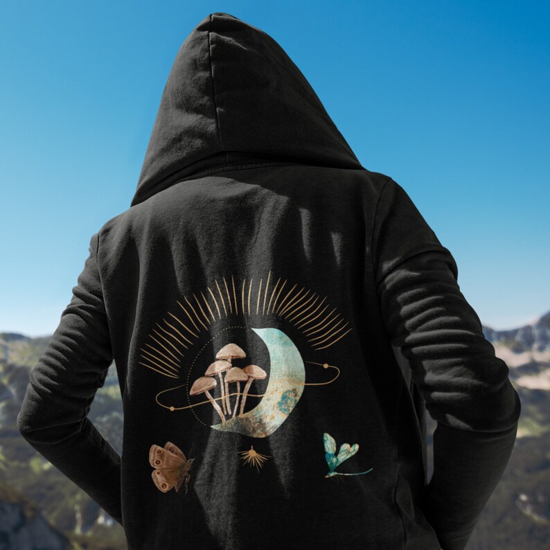 May include: Black hooded sweatshirt with a celestial design on the back. The design features a crescent moon, mushrooms, a butterfly, and a dragonfly. The artwork is in shades of brown, teal, and gold.