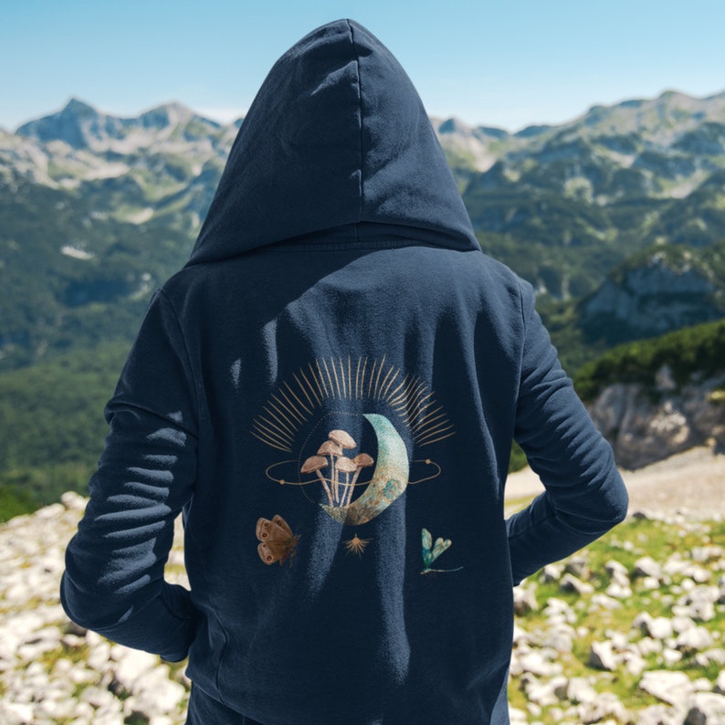 May include: Navy blue hooded sweatshirt with a celestial design on the back. The design features a crescent moon, mushrooms, butterflies, and stars in gold, brown, and green. The background is a mountain range.