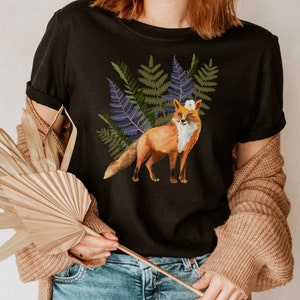 May include: Black t-shirt with a watercolour illustration of a fox wearing a flower crown, set against green and purple ferns. The fox has orange fur and a white chest. The t-shirt is a classic crew neck.