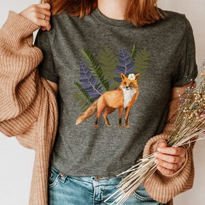 May include: Dark grey t-shirt with a watercolour illustration of a fox wearing a white flower crown, set against green and purple ferns. The fox has orange fur and a white chest. A nature-themed graphic tee.