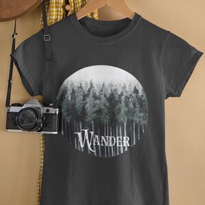 May include: Dark gray t-shirt with a circular graphic of a forest scene in shades of green and gray. The word "Wander" is printed in white across the bottom of the graphic. The shirt is hanging on a wooden hanger.