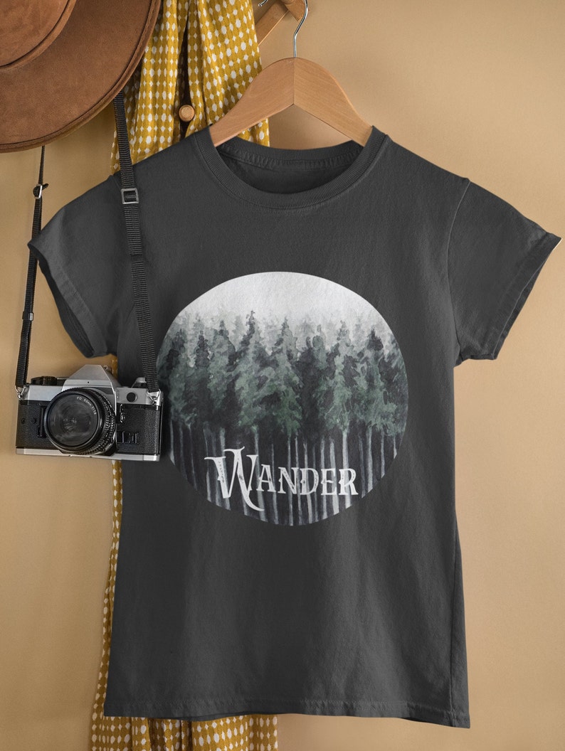May include: Dark gray t-shirt with a circular graphic of a forest scene in shades of green and gray. The word "Wander" is printed in white across the bottom of the graphic. The shirt is hanging on a wooden hanger.