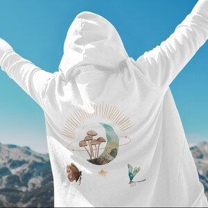 May include: White hooded sweatshirt with a nature-themed design on the back. The design includes a crescent moon, mushrooms, a butterfly, and a dragonfly. The background is a blue sky and mountains.