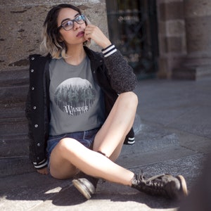 May include: A gray t-shirt featuring a circular forest graphic with the word "WANDER" in a stylized font. The shirt is paired with denim shorts and a black jacket with white striped cuffs. The overall aesthetic is casual and outdoorsy.