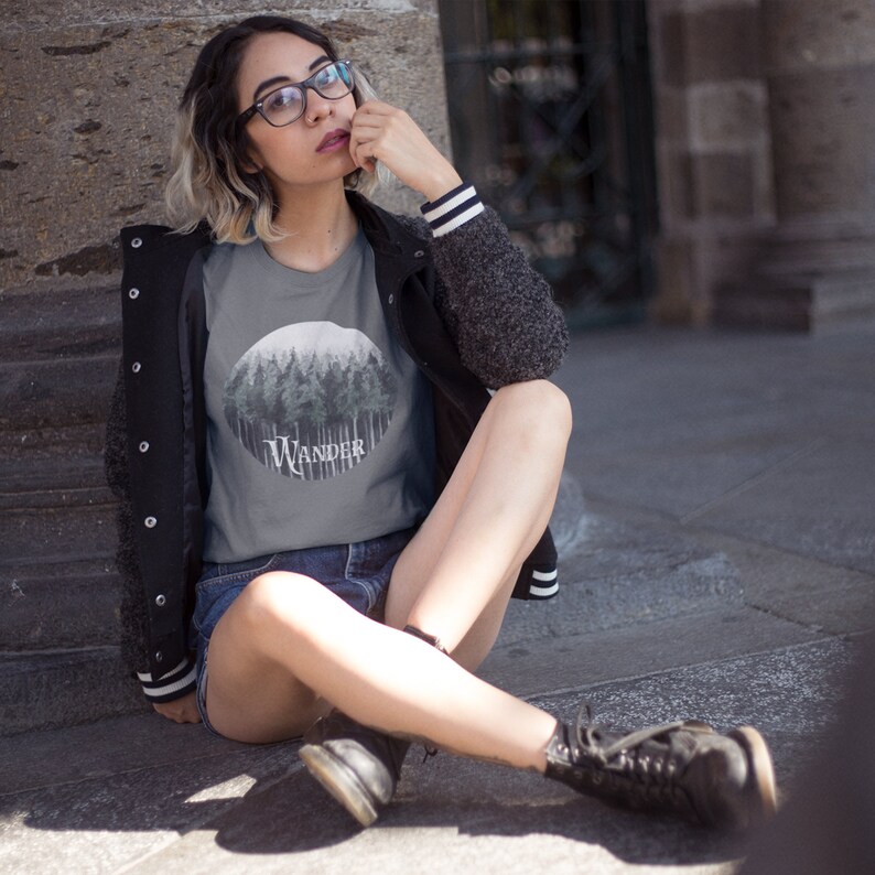 May include: A gray t-shirt featuring a circular forest graphic with the word "WANDER" in a stylized font. The shirt is paired with denim shorts and a black jacket with white striped cuffs. The overall aesthetic is casual and outdoorsy.