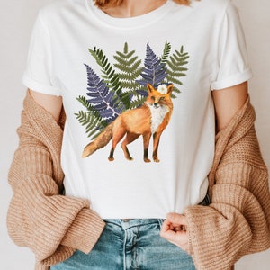May include: White t-shirt with a watercolour illustration of a fox. The fox has orange fur, a white chest, and a flower on its head, set against green and purple ferns. A casual, everyday wear item.