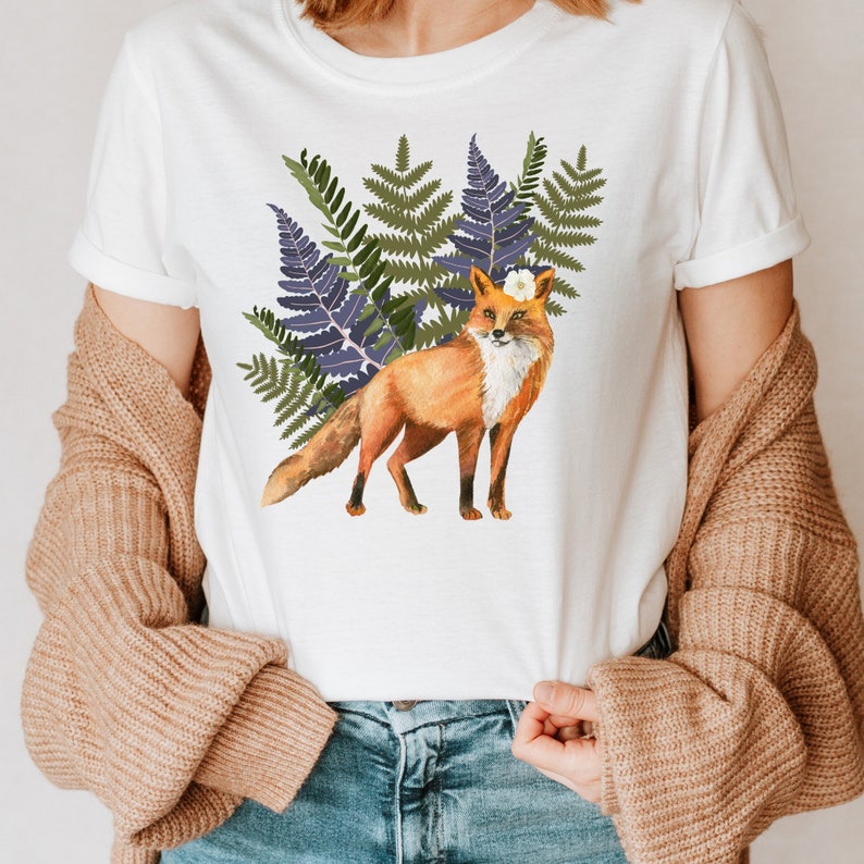 May include: White t-shirt with a watercolour illustration of a fox. The fox has orange fur, a white chest, and a flower on its head, set against green and purple ferns. A casual, everyday wear item.