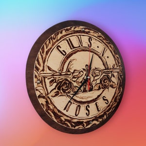May include: A wooden wall clock with a Guns N' Roses logo burned into the face. The clock has black hands and a brown wooden frame.