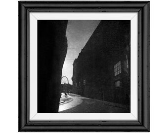 May include: Black and white framed print of a street scene. The image features a curved road, buildings, and a Ferris wheel in the distance. The frame is black with a white mat.