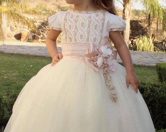May include: A young girl wearing a white tulle dress with a pink sash and a floral headband. The dress has a lace bodice and a tulle skirt. The girl is standing in a garden.