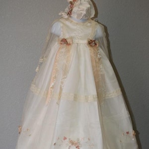 Puede incluir: Vestido de bautizo de color marfil con gorro a juego. El vestido presenta una falda en capas, delicados ribetes de encaje y detalles florales bordados. Cintas y lazos color melocotón añaden un toque de color.