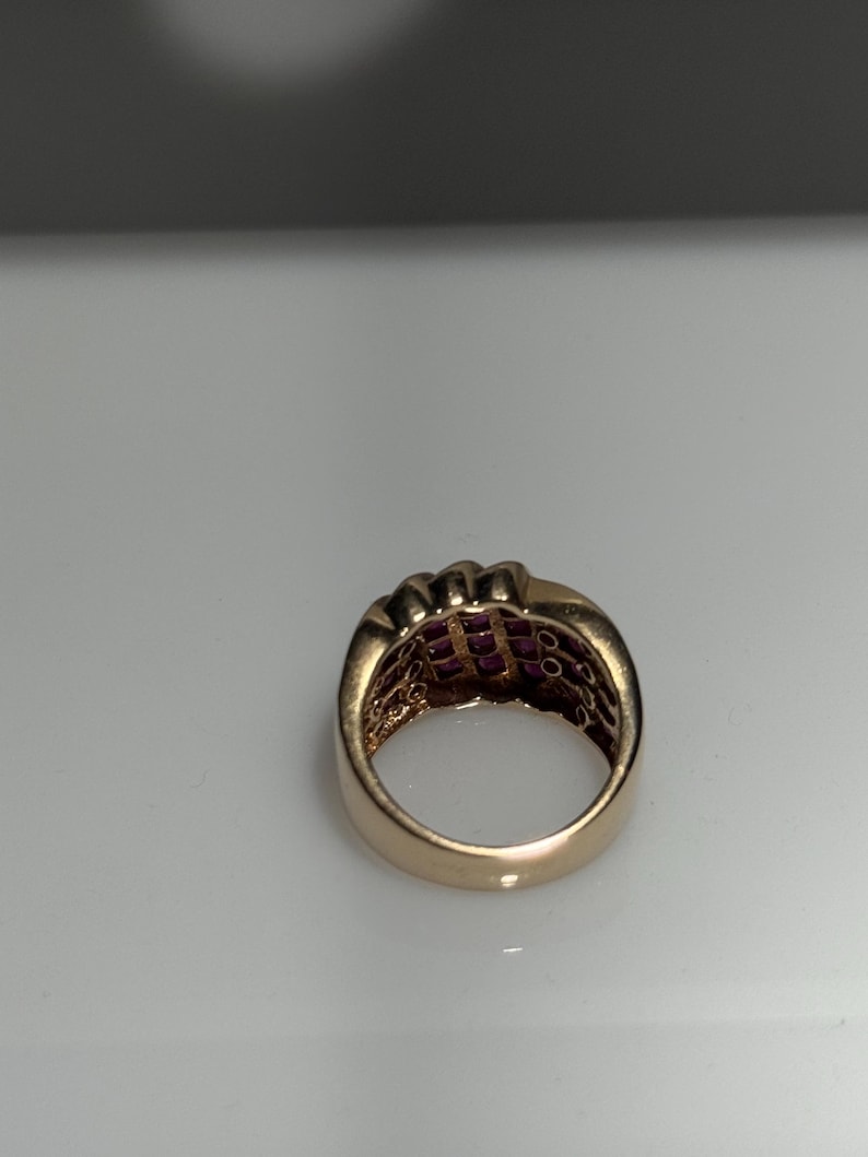 K&ouml;nnte beinhalten: Ein goldfarbener Ring mit mehreren Reihen kleiner, quadratisch geschliffener, roter Edelsteine. Der Ring hat ein breites Band und ein klassisches Design. Die Edelsteine sind eng aneinander gesetzt und bilden eine durchgehende, lebendige Oberfl&auml;che.