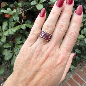 K&ouml;nnte beinhalten: Ein goldener Ring mit einer Reihe kleiner, roter Edelsteine. Der Ring befindet sich an einer Hand mit rotem Nagellack, vor dem Hintergrund gr&uuml;ner Bl&auml;tter.