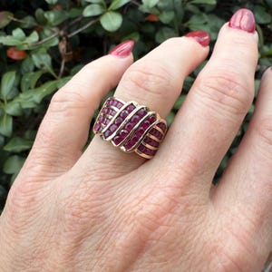 K&ouml;nnte beinhalten: Ein goldener Ring mit einer Reihe paralleler B&auml;nder, die jeweils mit kleinen, rechteckigen, tiefroten Edelsteinen besetzt sind. Der Ring befindet sich an einer Hand mit rotem Nagellack, vor gr&uuml;nem Laub.